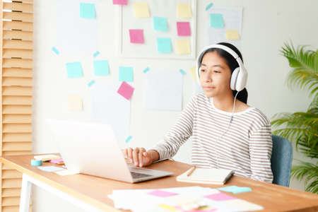 Happy Asian girl video call online via the internet tutor on a computer laptop with headphone. Concept online learning at homeの写真素材