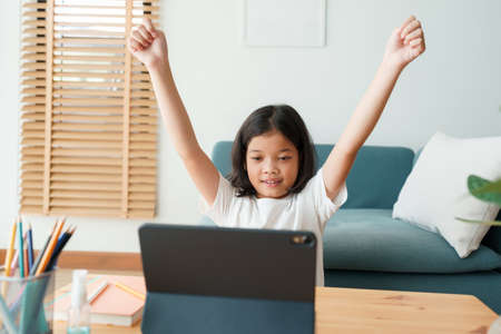 Happy Asian little girl raise hand is learning online with a tablet while sitting in living room in morning, Concept distance learning at homeの写真素材