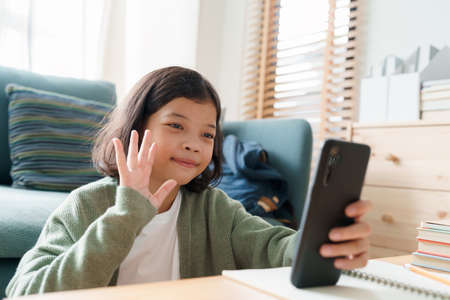 Happy Asian girl waving in greeting online tutor with a smartphone in the living room at home...