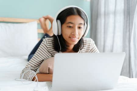 Asian girl using a laptop with Headphones while lying on the bed at home morning and sunlight, happy...