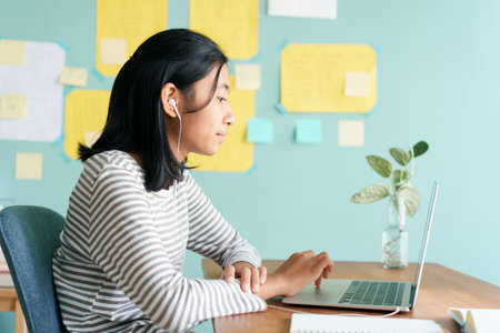 Happy Asian girl video call online via internet tutor on laptop computer with headphone while sitting on a table at home morning, Concept distance learning at homeの写真素材