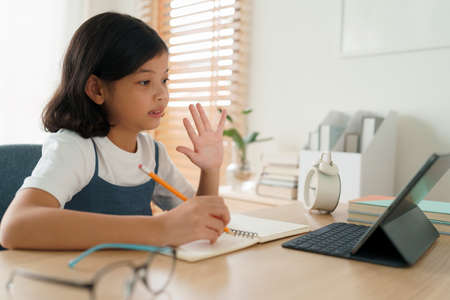 Asian little girl short hair waving in greeting online tutor with tablet while sitting at a table in...