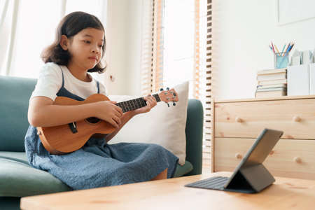Asian little girl is practicing ukulele playing and learn with a teacher online via internet on tablet digital at home morningの写真素材