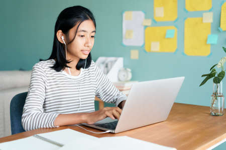 Happy Asian girl video call online via internet tutor on laptop computer with headphone while sitting on a table at home morning, Concept distance learning at homeの写真素材