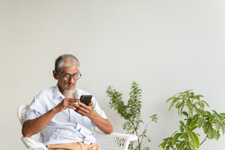 Pleasant senior older men resting while sitting on chair, Asian elder man with grey hair using application on smartphone at home morningの写真素材