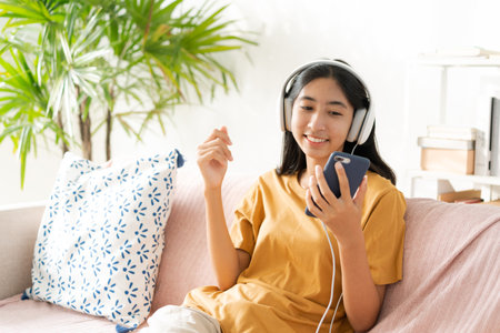 Asian woman listening to music on smartphone with headset enjoy. Asia woman using mobile phone happy smile while sitting on sofa at home morning lightの写真素材