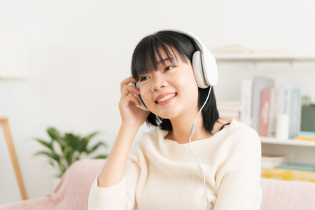 Asian teenager listening to music on tablet with headphone enjoy. Asia woman using computer happy smile while sitting on sofa at home morningの写真素材