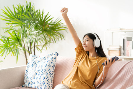 Asian teenager listening to music on smartphone with headset enjoy. raise hand, Asia woman using mobile phone happy smile relax at home morning lightの写真素材