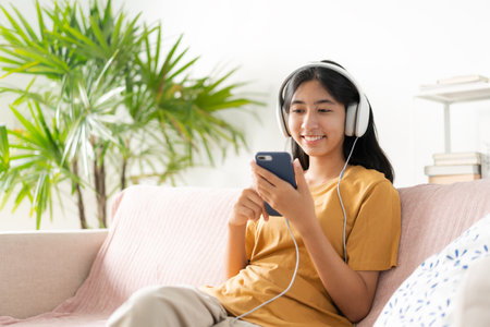 Asian teenager listen to music on smartphone with headphone enjoy. Asia woman using mobile phone happy smile  relax at home morning lightの写真素材