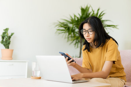 Asian woman wear glasses typing work on laptop. Asia student learn via internet with computer, using mobile phone,  while sitting on sofa in living room at homeの写真素材
