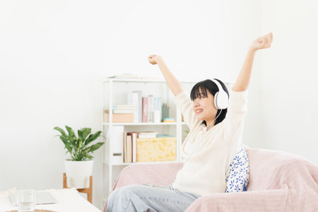 Asian teenager listen to music on smartphone with headphone enjoy. raise-up hand, Asia woman using mobile phone happy smile while sitting on sofa at home morningの写真素材