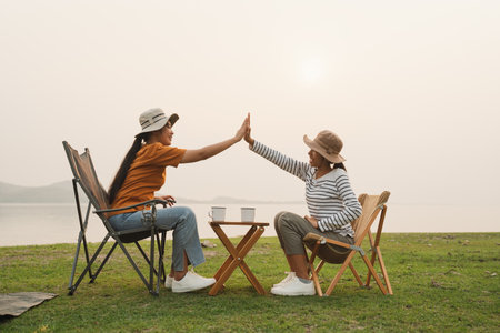 Asian two teenager high five gesture while sitting at camping sunset. Happy asia teen girl smile for celebrating together at campsite outdoor. Camping in vacation concept summer timeの写真素材