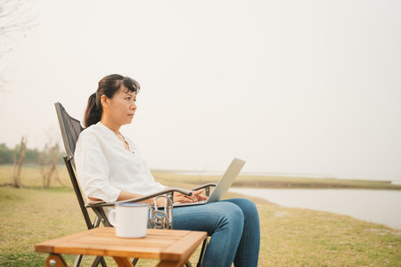 Asian woman typing on laptop while sitting at campsite outdoor. Asia women relax working using computer amidst meadow field and river.  Concept camping of lifestyle vacation and freedomの写真素材