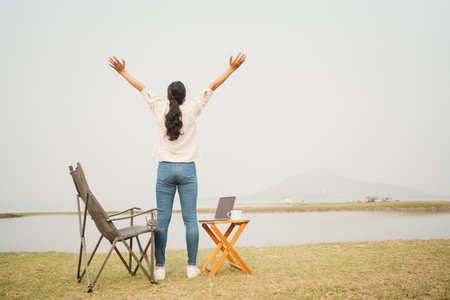 Back view. Asian woman raised up hand looking at river. Happy asia women relaxing after working campsite outdoor. Concept camping of lifestyle vacation and freedomの写真素材