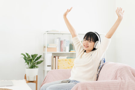 Asian teenager listen to music on smartphone with headphone enjoy. raise-up hand, Asia woman using mobile phone happy smile while sitting on sofa at home morningの写真素材