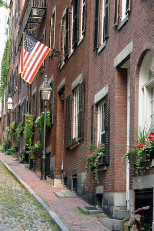 Walking up the cobblestone alley called Acorn Street lined with brick buildings, brick sidewalks old gas lanterns wooden shutters, flower boxes and an early american flagの写真素材