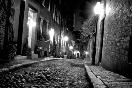 old 19th Century cobble stone road in Boston Massachusetts, lit only by the gas lamps revealing the shuttered windows and brightly lit doorways of the rowhouses on Acorn Streetの写真素材