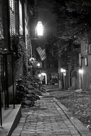 black and white night image of an old 19th Century cobble stone road in Boston Massachusetts, lit only by the gas lamps revealing the shuttered windows and brightly lit doorways of the rowhouses on Acorn Street, you can almost feel the night coming downの写真素材