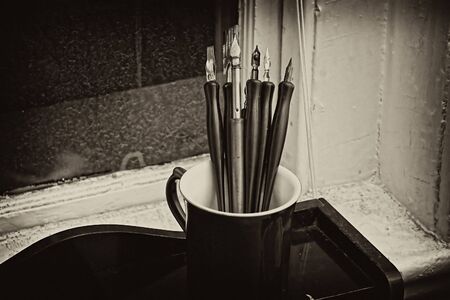 artists cup full of old dip pens sits on wooden desk in this toned imageの写真素材
