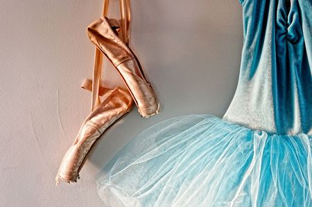 a blue velvet romantic tutu is hanging on a wall beside a worn pair of ballet pointe shoes, lit only by sunlight through windowの写真素材