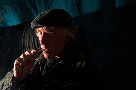 A dangerous looking man with an evil expression on his face is sitting in a dark room looking at viewer smoking pipe.の写真素材