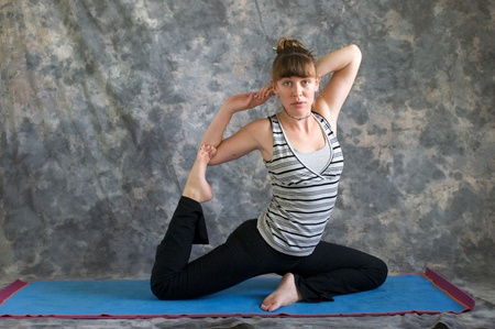 Young woman on yoga mat  doing Yoga posture Kapotasana or Pigeon Pose variation against a grey background in profile, facing right lit by diffused sunlight.の写真素材