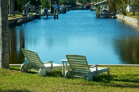 Two white outside lounge chairs are sitting by the water at the mouth of a canal in florida.の写真素材