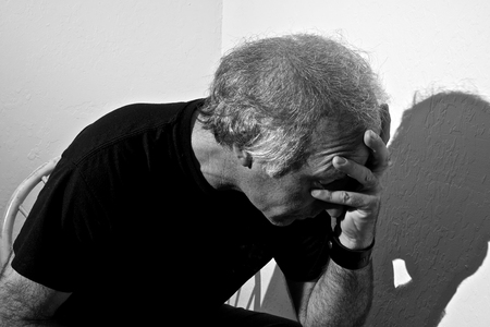 Portrait of a distressed older white man holding head sitting in corner of white room.の写真素材