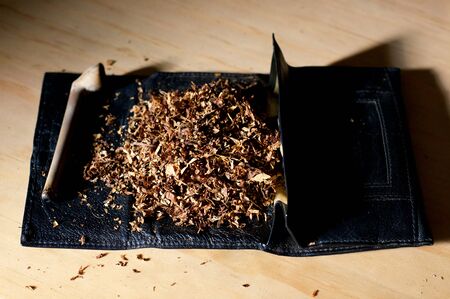 An old clay pipe is laid beside a pile of tobacco on black leather pouch. Lit by morning sunlight.の写真素材