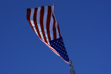 Looking up at American red, white, and blue flag on flagpole from directly below on windy day against clear blue sky.の写真素材