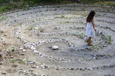 A young woman with long brown hair and white dress is walking away from the center of a maze or stone labyrinth.の写真素材