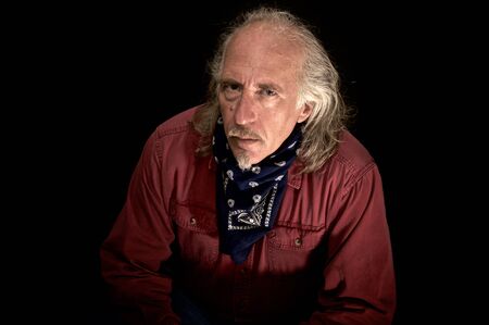 An old rugged looking cowboy with red shirt and blue bandana, long hair and mustache and goatee looking at camera with head cocked as listening.の写真素材