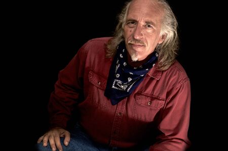 An old rugged looking cowboy with red shirt and blue bandana, long hair and mustache and goatee looking at camera smiling.の写真素材
