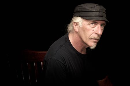 An older white biker male sitting in a dark room wearing black t shirt and leather cap, looking serious at viewer, three quarter view portrait.の写真素材