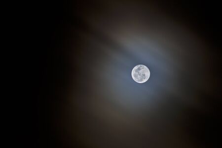 A bright full moon showing detail of surface with fast moving wispy clouds creating streaks around moonの写真素材