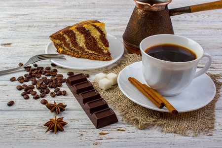 A cup of coffee on sacking with tasty chocolate cake on a plate. Cinnamon sticks, sugar, coffee beans, a chocolate bar, anice on bright wooden tableの写真素材