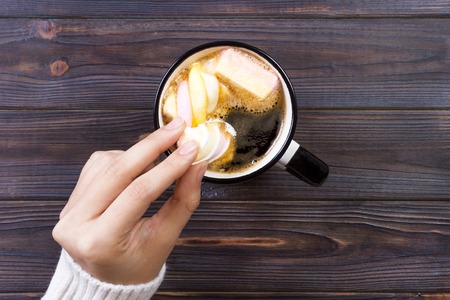 female hand holding cup of hot cocoa or chocolate with marshmallow on wooden table from above.の写真素材