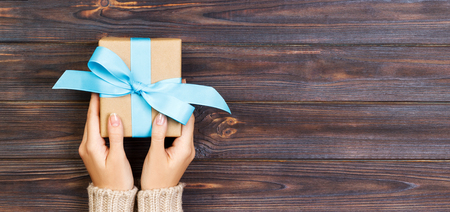 Woman hands give wrapped valentine or other holiday handmade present in paper with blue ribbon. Present box, decoration of gift on white wooden table, top view with copy space. Banner.の写真素材