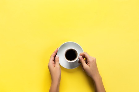 Minimalistic style woman hand holding a cup of coffee on yellow background. Flat lay, top view.の写真素材
