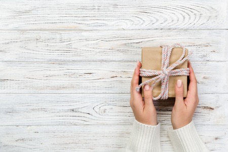 Woman hands give wrapped valentine or other holiday handmade present in paper with pink ribbon. Present box, decoration of gift on white wooden table, top view with copy space.の写真素材