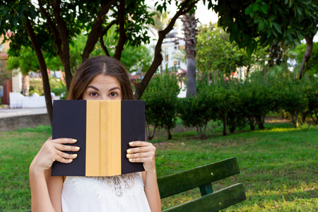 Young cute girl is covering her face with a book. Surprised and shocked woman. Emotional girl holding a book in her hands on the bench in the park.の写真素材