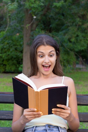 Surprised young woman with widely open yeas and mouth is reading a book outdoors.の写真素材