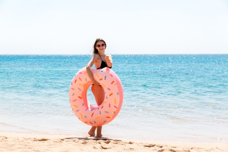 woman calls to swim in the sea and waves her hand. Girl relaxing with donut on the beach and playing...