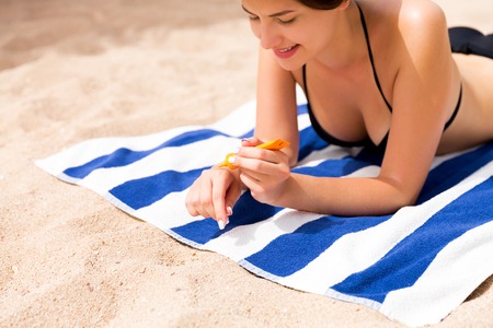 Beautiful lady is sunbathing on the towel on the sand at the beach and protects her hands with sunblock.の写真素材