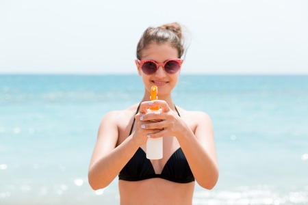 Young woman is holding a spray of sun cream for body at the camera at the beach.