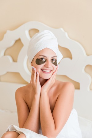 Beautiful young woman with under eye patches in bathrobe sitting in bed. Happy girl taking care of herself. Beauty skincare and wellness morning concept.の写真素材