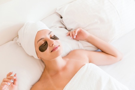 Beautiful young woman with under eye patches in bathrobe lying in bed. Happy girl taking care of herself. Beauty skincare and wellness morning concept.の写真素材