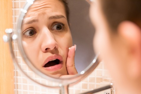 Young girl in front of a bathroom mirror putting cream on a red pimple. Beauty skincare and wellness morning concept.の写真素材