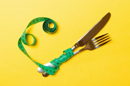 Crossed fork and knife are wrapped in measuring tape on yellow background. Concept of diet for...