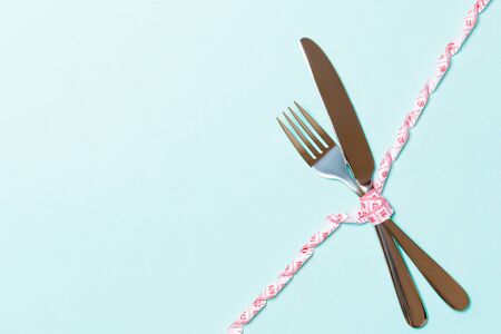 Diet and healthy eating concept with crossed fork and knife and curled measuring tape on blue...